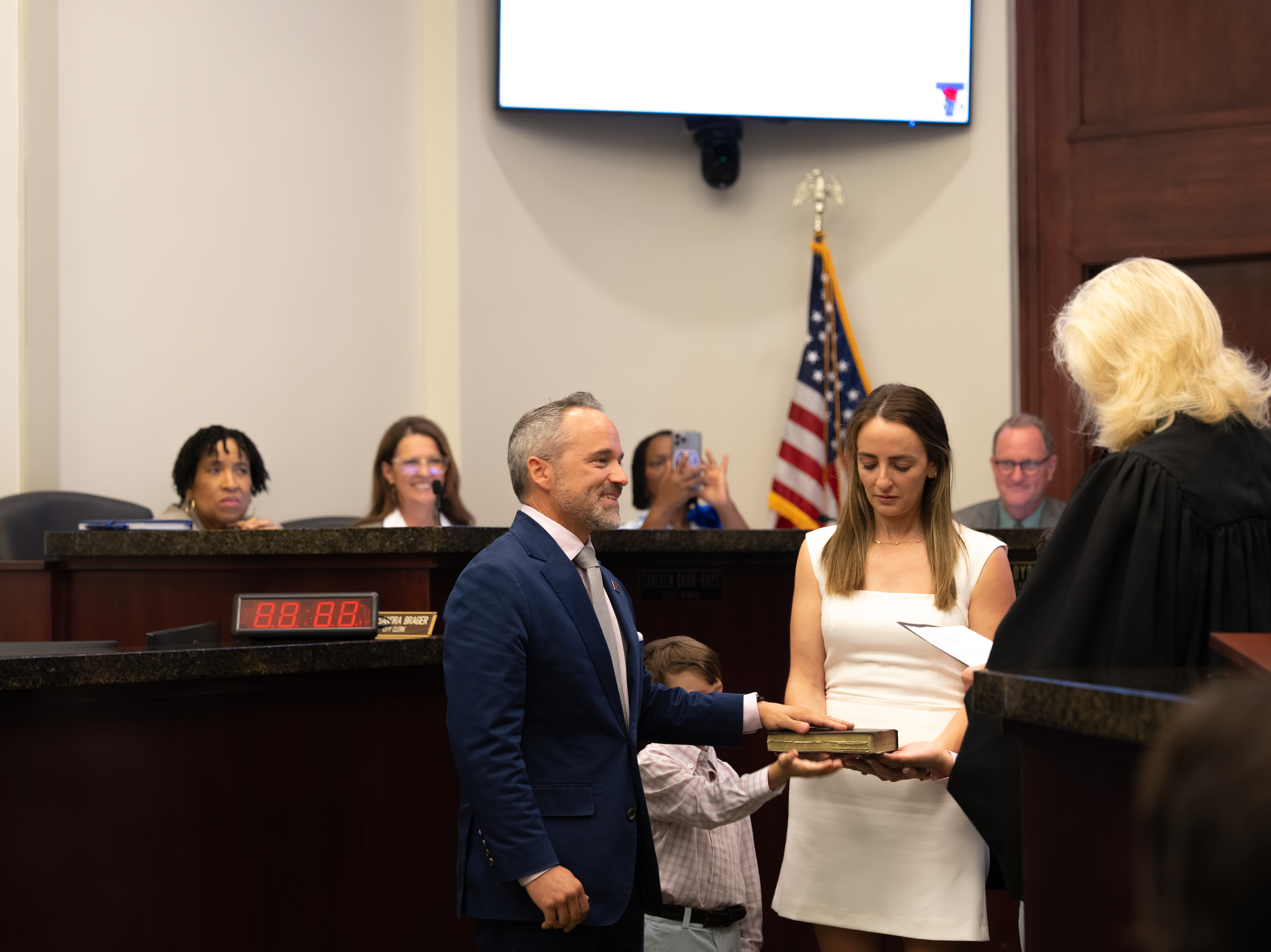 Man in a suit taking an oath from a judge in a courtroom while a woman in a white dress holds the book and a child stands nearby.