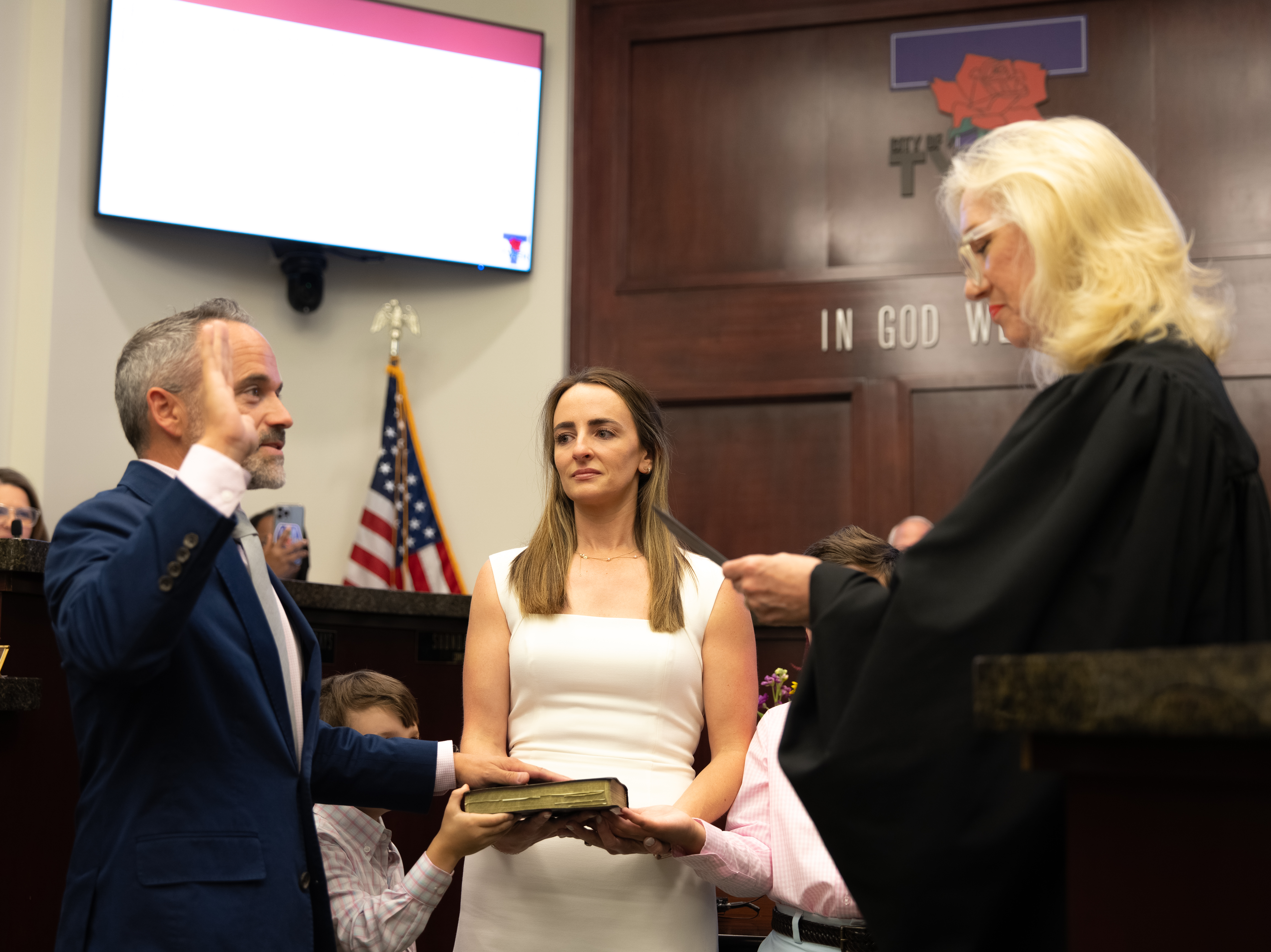 Man in suit raising his right hand to take an oath in a courtroom while a woman holds a book and a judge administers the oath.