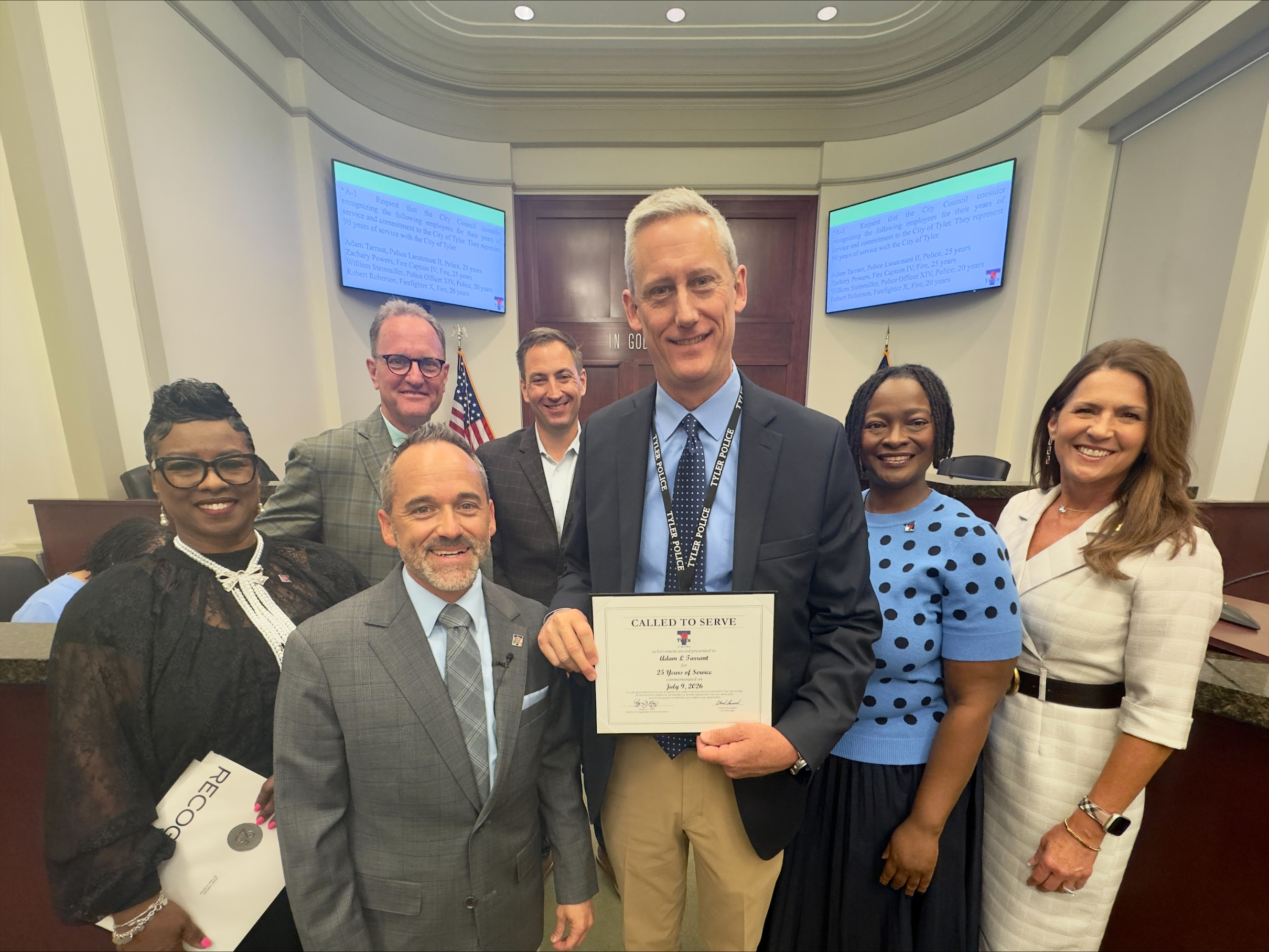 Seven people pose for a group photo inside the Tyler City Council chambers. A man in the center holds a “Called to Serve” certificate recognizing 25 years of service.