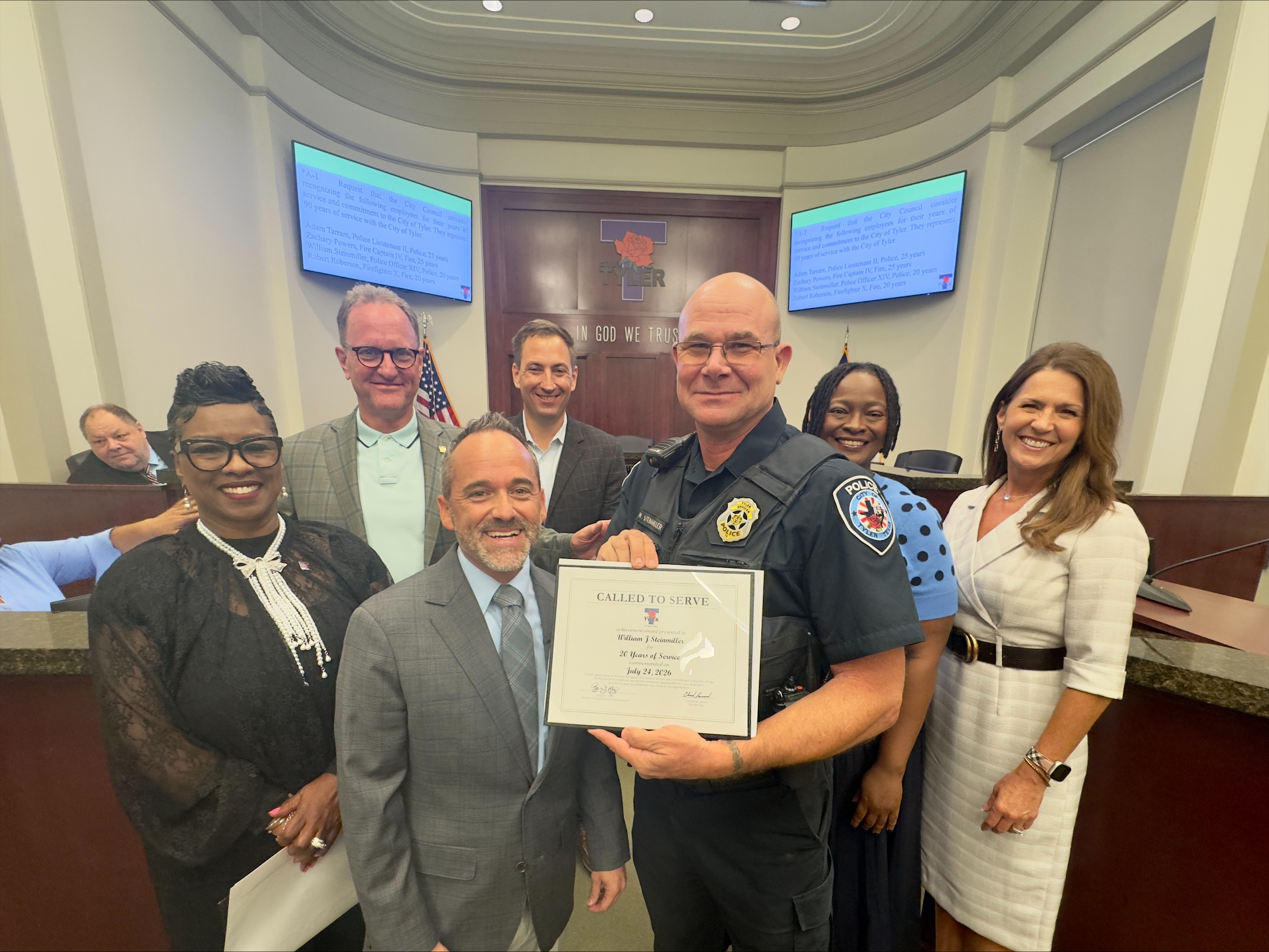 Seven people pose for a group photo inside the Tyler City Council chambers. A Tyler Police officer in the center holds a “Called to Serve” certificate recognizing 20 years of service.