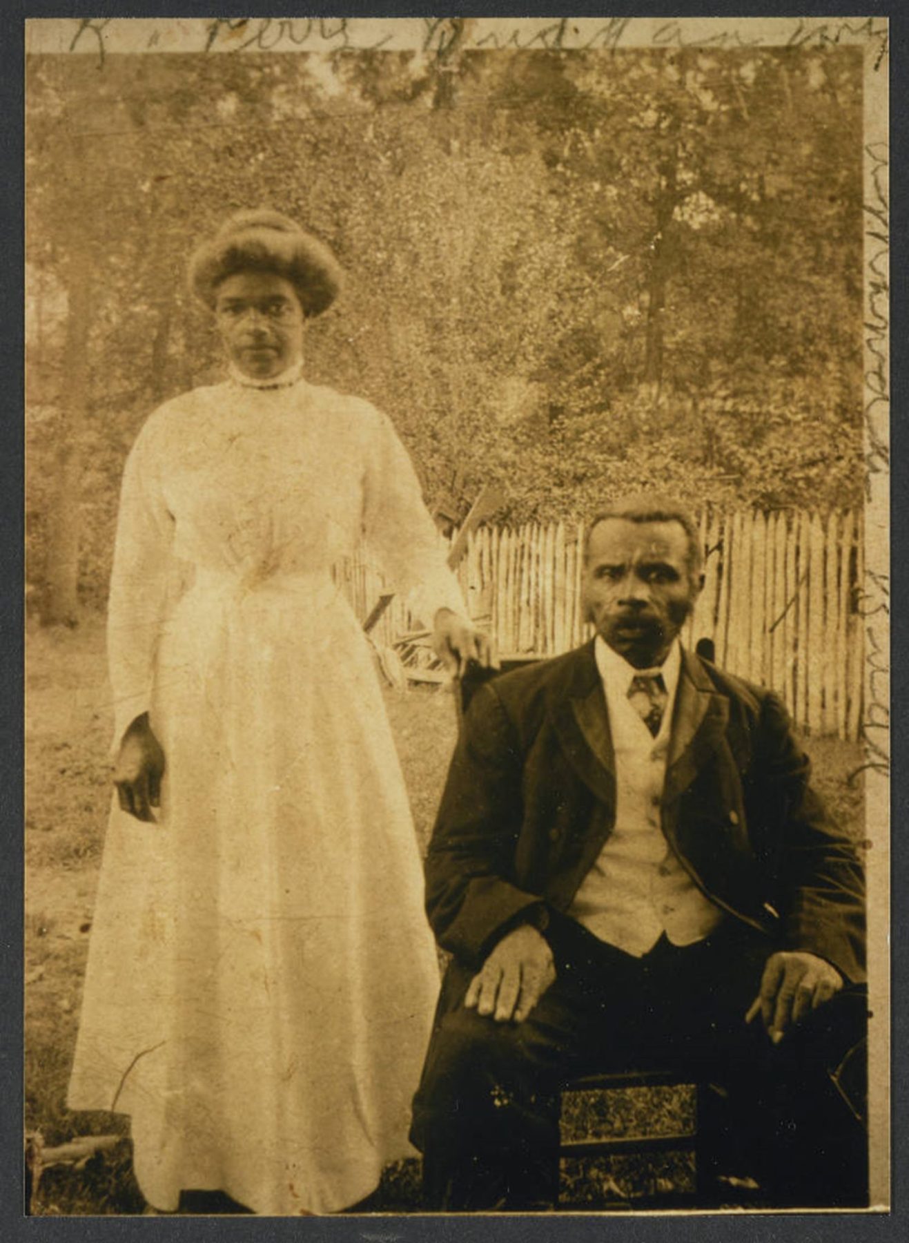 A woman stands beside a seated man outdoors in front of a wooden fence and trees. Both wear early 20th-century formal clothing and look directly at the camera.