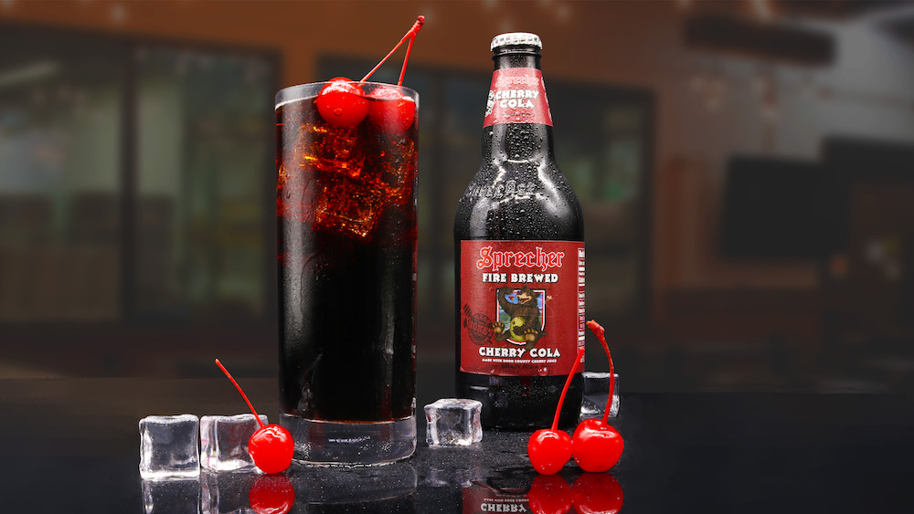 A bottle and glass of cherry cola on a bar surrounded by ice and maraschino cherries