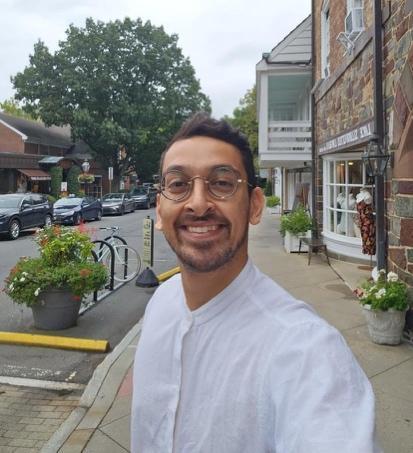 The image shows a picturesque street scene with a row of charming shops and a few parked cars, surrounded by greenery and quaint buildings.AI-generated content may be incorrect.