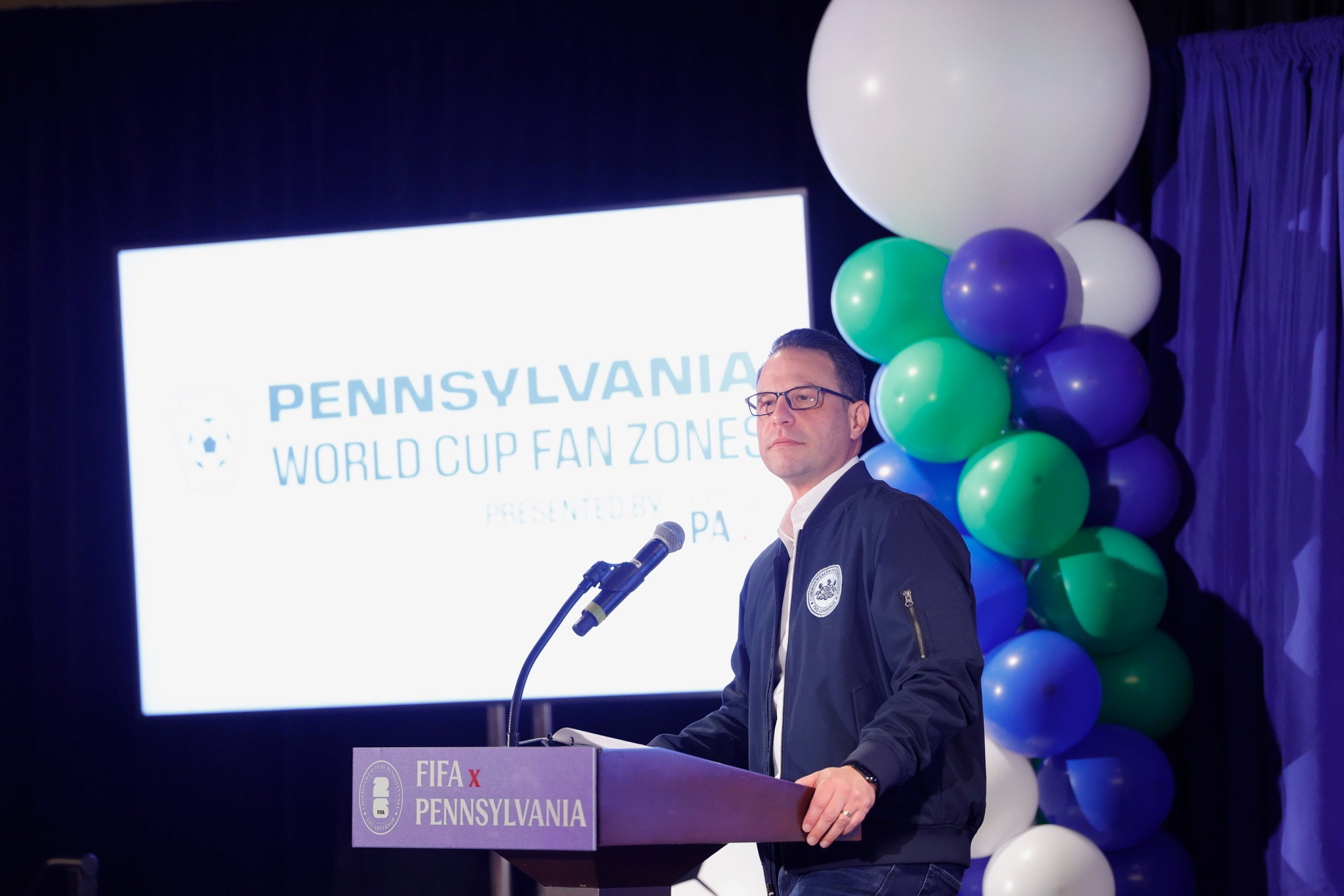Man speaking on a stage at a podium with a screen behind him that reads Pennsylvania World Cup Fan Zone